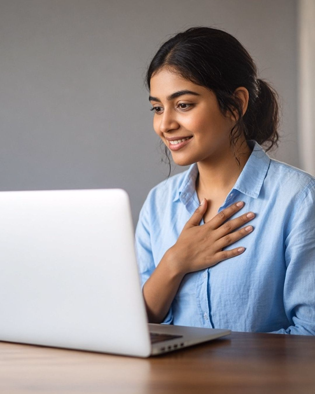 A relieved young graduate smiles at her laptop with a hand on her chest
